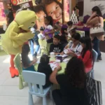 A group of children are sitting around a table with a mascot.